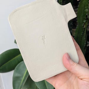 Puede incluir: Una bandeja rectangular de cer&aacute;mica color blanco hueso con acabado mate y una peque&ntilde;a asa en el lado derecho. El centro de la bandeja presenta una firma o logotipo en relieve que dice "mumu" en una fuente cursiva min&uacute;scula.