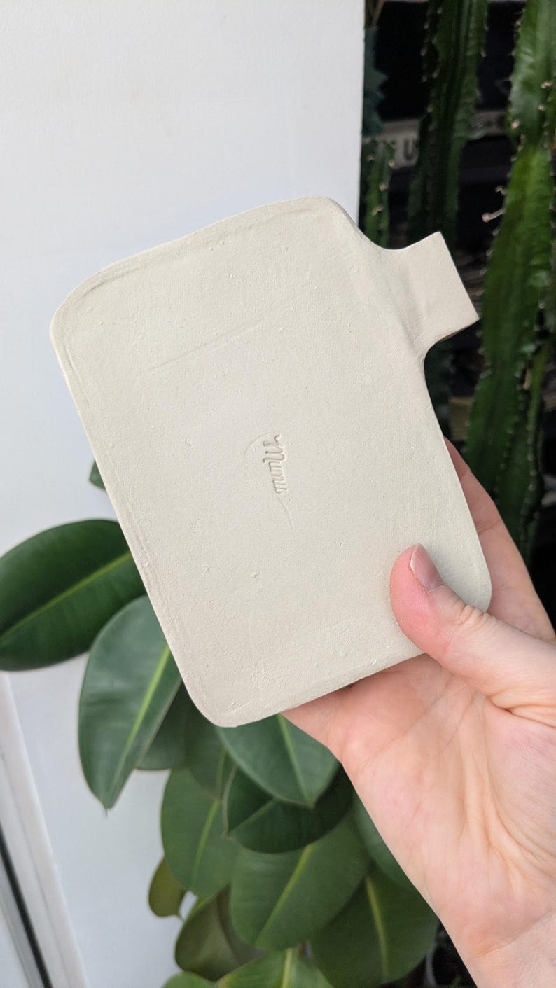Puede incluir: Una bandeja rectangular de cer&aacute;mica color blanco hueso con acabado mate y una peque&ntilde;a asa en el lado derecho. El centro de la bandeja presenta una firma o logotipo en relieve que dice "mumu" en una fuente cursiva min&uacute;scula.