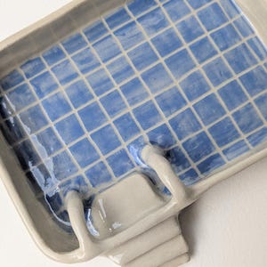 Puede incluir: Una bandeja rectangular de cer&aacute;mica con acabado brillante, que presenta un patr&oacute;n de cuadr&iacute;cula azul y blanco en el interior que recuerda a una piscina. La bandeja incluye una escalera de cer&aacute;mica beige esculpida en un lado, que se extiende hacia el interior.
