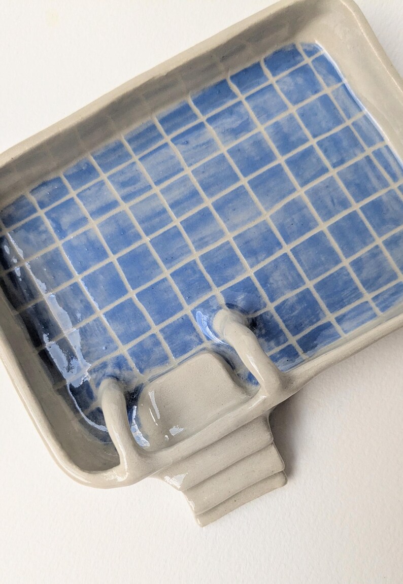 Puede incluir: Una bandeja rectangular de cer&aacute;mica con acabado brillante, que presenta un patr&oacute;n de cuadr&iacute;cula azul y blanco en el interior que recuerda a una piscina. La bandeja incluye una escalera de cer&aacute;mica beige esculpida en un lado, que se extiende hacia el interior.