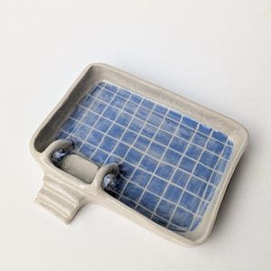 Puede incluir: Una jabonera rectangular de cer&aacute;mica con forma de piscina, con un borde gris claro y un patr&oacute;n interior de azulejos azules. La pieza presenta una peque&ntilde;a escalera de tres pelda&ntilde;os en un lado, que imita una escalera de piscina.