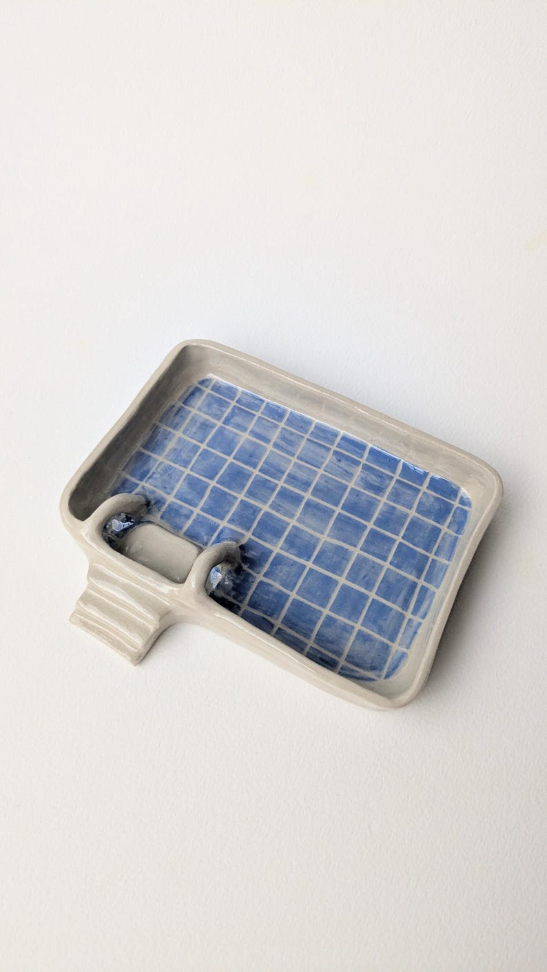 Puede incluir: Una jabonera rectangular de cer&aacute;mica con forma de piscina, con un borde gris claro y un patr&oacute;n interior de azulejos azules. La pieza presenta una peque&ntilde;a escalera de tres pelda&ntilde;os en un lado, que imita una escalera de piscina.