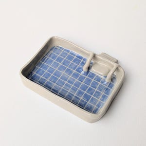 Puede incluir: Una bandeja rectangular de cer&aacute;mica con borde exterior gris claro y un interior azul brillante con un patr&oacute;n de cuadr&iacute;cula blanca. La bandeja incluye una peque&ntilde;a estructura de trampol&iacute;n de cer&aacute;mica gris unida a un lado.