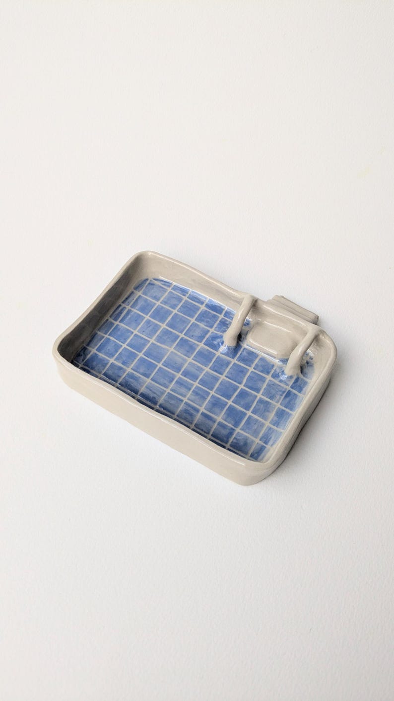 Puede incluir: Una bandeja rectangular de cer&aacute;mica con borde exterior gris claro y un interior azul brillante con un patr&oacute;n de cuadr&iacute;cula blanca. La bandeja incluye una peque&ntilde;a estructura de trampol&iacute;n de cer&aacute;mica gris unida a un lado.