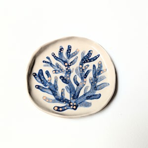 May include: A small, round ceramic plate featuring a hand-painted coral reef design. The coral is a deep blue with peach-coloured dots, set against a cream background. The plate has a slightly irregular edge.