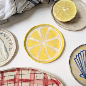 May include: A selection of handmade ceramic plates with diverse designs. One plate showcases a yellow lemon slice, another has a red plaid pattern, and others display seashell and oyster motifs. A halved lemon and a blue and white checkered cloth are also present.