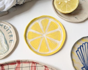 May include: A collection of handmade ceramic plates with various designs. One plate features a yellow lemon slice, another has a red plaid pattern, and others display seashell and oyster motifs. A halved lemon and a blue and white checkered cloth are also present.