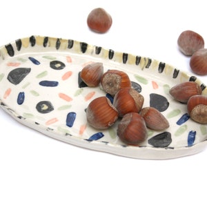 May include: A white ceramic oval serving tray with a black and yellow striped rim and a colorful abstract pattern. The tray is filled with brown hazelnuts.