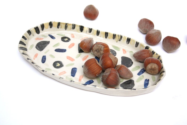 May include: A white ceramic oval serving tray with a black and yellow striped rim and a colorful abstract pattern. The tray is filled with brown hazelnuts.