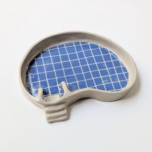 May include: A kidney-shaped ceramic dish with a glossy light grey exterior and a blue interior featuring a white grid pattern that resembles a swimming pool. The dish includes a small, attached three-step staircase detail on one side.
