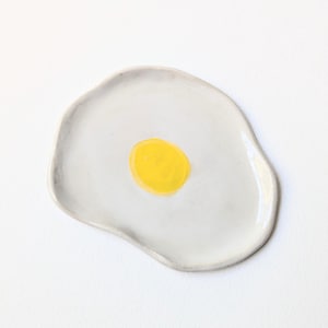 May include: A small, irregular, hand-formed ceramic dish with a glossy white finish and a single, centered, bright yellow circular painted accent, resembling the appearance of a fried egg on a plate, with smooth, rounded, organic edges.