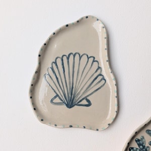 Peut inclure: Un petit plat en céramique beige de forme irrégulière et organique. Le centre présente une illustration peinte à la main d'un coquillage Saint-Jacques bleu foncé. Le bord extérieur est orné de petits points alternés bleu foncé et bleu sarcelle.