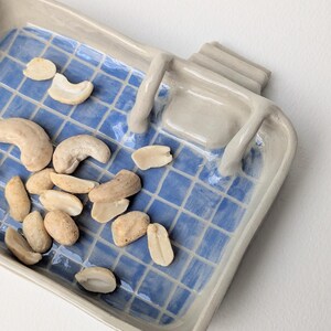 Puede incluir: Una bandeja rectangular de cer&aacute;mica con un patr&oacute;n de cuadr&iacute;cula azul brillante sobre una base blanca, con bordes beige elevados y una peque&ntilde;a estructura en forma de escalera en un lado. La bandeja contiene nueces de la India curvas.