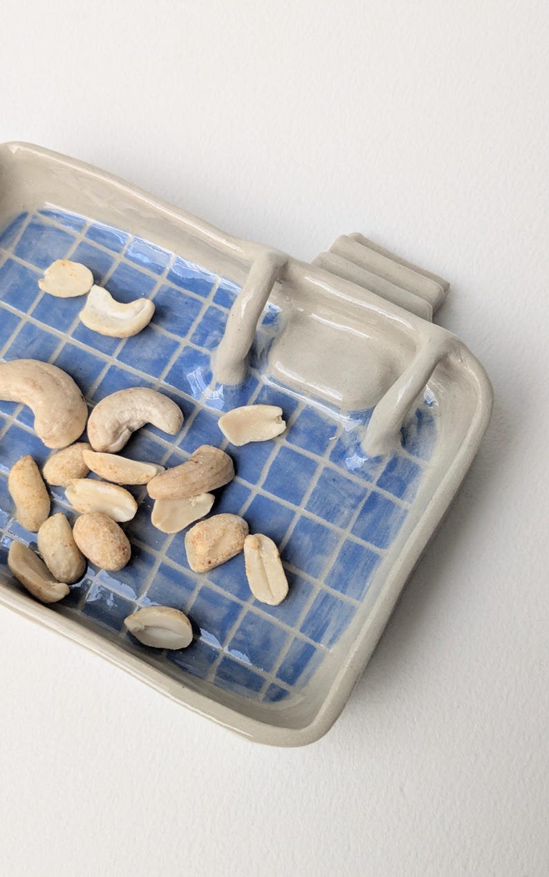 Puede incluir: Una bandeja rectangular de cer&aacute;mica con un patr&oacute;n de cuadr&iacute;cula azul brillante sobre una base blanca, con bordes beige elevados y una peque&ntilde;a estructura en forma de escalera en un lado. La bandeja contiene nueces de la India curvas.