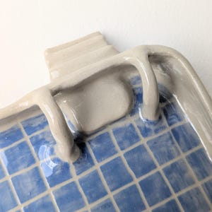 Puede incluir: Una bandeja de cer&aacute;mica con acabado brillante que presenta un patr&oacute;n de cuadr&iacute;cula azul y blanco que recuerda a una piscina. La bandeja tiene un borde elevado de color gris claro con un elemento esculpido en forma de escalera en un lado.