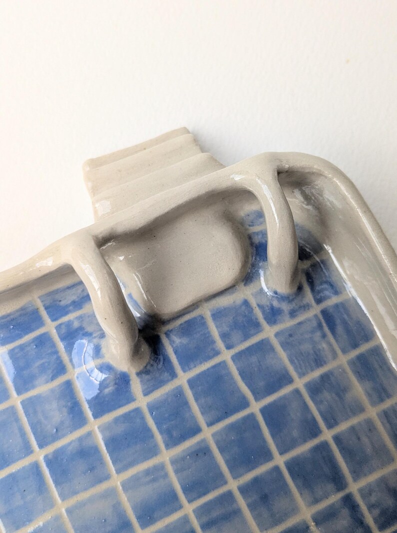 Puede incluir: Una bandeja de cer&aacute;mica con acabado brillante que presenta un patr&oacute;n de cuadr&iacute;cula azul y blanco que recuerda a una piscina. La bandeja tiene un borde elevado de color gris claro con un elemento esculpido en forma de escalera en un lado.