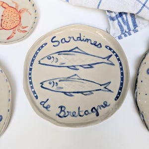 Puede incluir: Un plato de cerámica blanco con un borde azul e ilustraciones azules y blancas de sardinas y el texto "Sardines de Bretagne".