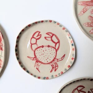 Puede incluir: Un conjunto de platos de cerámica con un diseño de cangrejo pintado a mano. El cangrejo es rojo, con un cuerpo punteado y contornos definidos. Los platos tienen un fondo crema y un borde punteado en rojo, azul y verde.