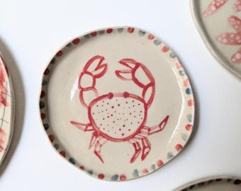 May include: A set of ceramic plates with a hand-painted crab design. The crab is red, with a dotted body and outlined features. The plates have a cream-colored background and a dotted border in red, blue, and green.