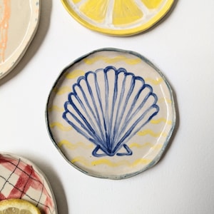 Puede incluir: Una colección de platos decorativos de cerámica. Un plato presenta un diseño de concha azul sobre un fondo crema con líneas onduladas amarillas. Otros platos muestran una rodaja de limón y un patrón de cuadros rojos y blancos.