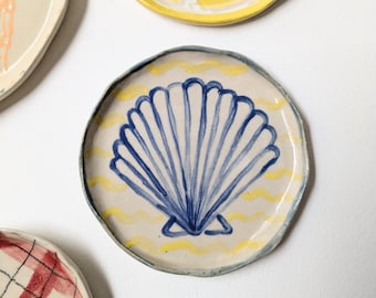 May include: A collection of decorative ceramic plates. One plate features a blue seashell design on a cream background with yellow wavy lines. Other plates display a lemon slice, and a red and white plaid pattern.