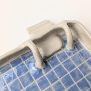 Puede incluir: Una bandeja rectangular de cer&aacute;mica con acabado brillante, que presenta un patr&oacute;n de cuadr&iacute;cula azul pintado a mano en la superficie interior. La bandeja tiene un borde elevado de color beige claro y dos peque&ntilde;as asas curvas.