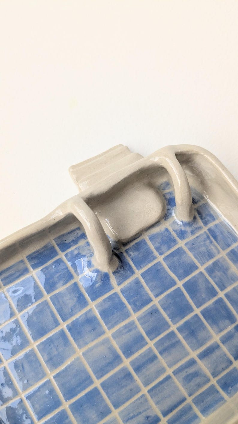 Puede incluir: Una bandeja rectangular de cer&aacute;mica con acabado brillante, que presenta un patr&oacute;n de cuadr&iacute;cula azul pintado a mano en la superficie interior. La bandeja tiene un borde elevado de color beige claro y dos peque&ntilde;as asas curvas.