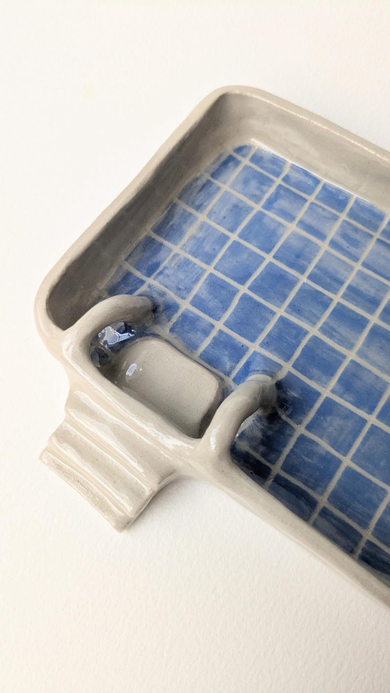 Puede incluir: Una bandeja rectangular de cer&aacute;mica con acabado beige brillante y un patr&oacute;n de cuadr&iacute;cula azul en la base interior. La bandeja presenta un elemento de escalera de cer&aacute;mica esculpido en un lado, que se extiende desde el borde hacia el interior.