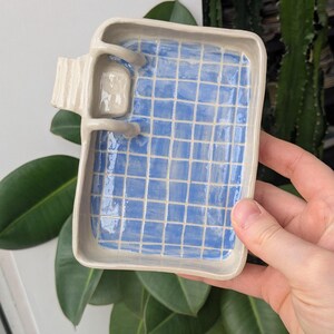 Puede incluir: Una bandeja rectangular de cer&aacute;mica con borde beige claro y un interior azul brillante con un patr&oacute;n de cuadr&iacute;cula blanca que recuerda a una piscina. Una peque&ntilde;a escalera de cer&aacute;mica esculpida est&aacute; unida a un lado de la bandeja.