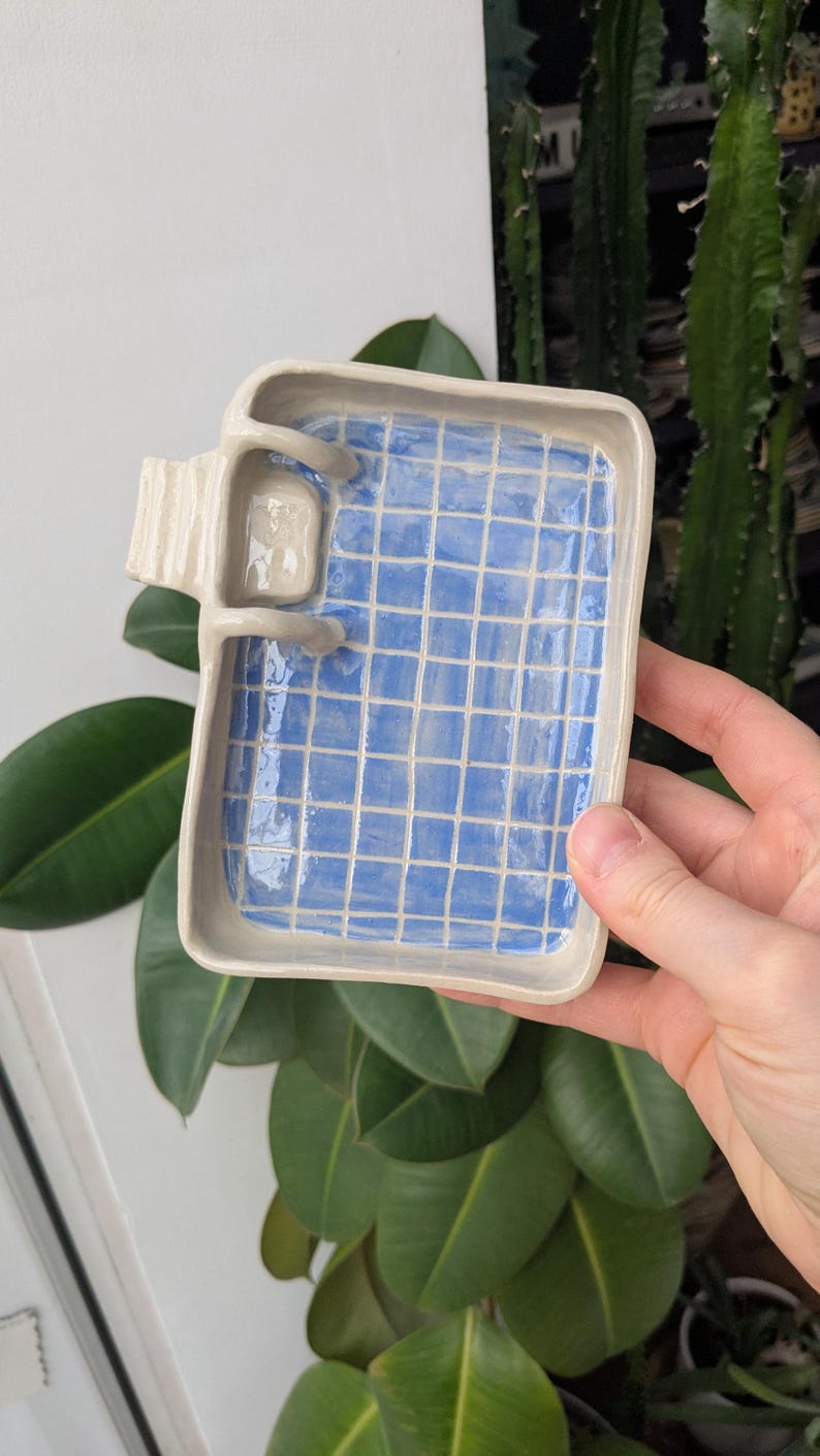 Puede incluir: Una bandeja rectangular de cer&aacute;mica con borde beige claro y un interior azul brillante con un patr&oacute;n de cuadr&iacute;cula blanca que recuerda a una piscina. Una peque&ntilde;a escalera de cer&aacute;mica esculpida est&aacute; unida a un lado de la bandeja.