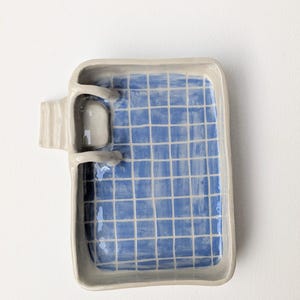 Puede incluir: Una bandeja rectangular de cer&aacute;mica con borde gris claro y un interior azul con un patr&oacute;n de cuadr&iacute;cula blanca. Una peque&ntilde;a escalera de cer&aacute;mica tridimensional est&aacute; unida al lado izquierdo de la bandeja, sobresaliendo del borde.