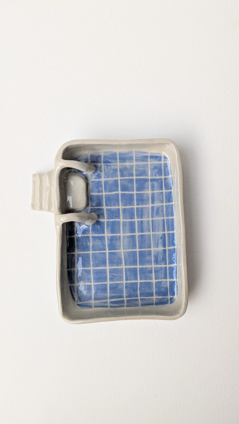 Puede incluir: Una bandeja rectangular de cer&aacute;mica con borde gris claro y un interior azul con un patr&oacute;n de cuadr&iacute;cula blanca. Una peque&ntilde;a escalera de cer&aacute;mica tridimensional est&aacute; unida al lado izquierdo de la bandeja, sobresaliendo del borde.