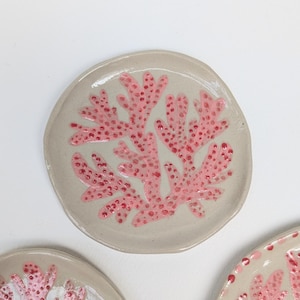 May include: A small, round ceramic plate featuring a coral reef design. The plate is a light beige colour with a pink coral pattern. The coral is dotted with darker pink spots. Part of a set of plates.