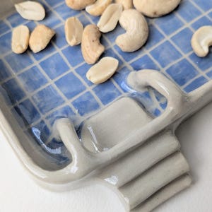 Puede incluir: Una bandeja rectangular de cer&aacute;mica con un patr&oacute;n de cuadr&iacute;cula azul y blanco brillante en la superficie interior. La bandeja presenta un borde elevado gris claro y un peque&ntilde;o elemento de escalera de tres pelda&ntilde;os en un lado, con anacardos.
