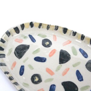 May include: A white ceramic oval serving dish with a black and yellow striped rim. The dish is decorated with hand-painted black, blue, green, and pink abstract shapes.