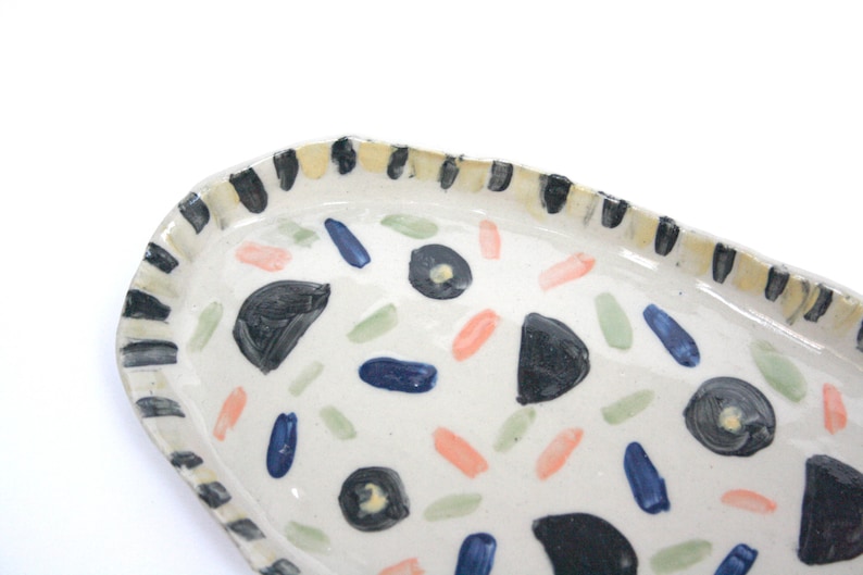 May include: A white ceramic oval serving dish with a black and yellow striped rim. The dish is decorated with hand-painted black, blue, green, and pink abstract shapes.