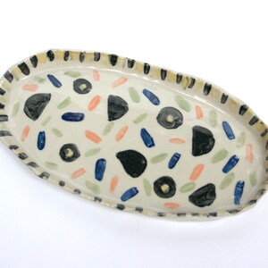 May include: A white oval ceramic serving dish with a black and white striped rim. The dish is decorated with black, blue, green, and orange abstract shapes.
