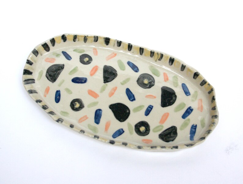 May include: A white oval ceramic serving dish with a black and white striped rim. The dish is decorated with black, blue, green, and orange abstract shapes.