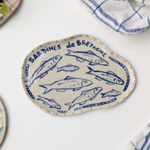 Könnte beinhalten: Ein unregelmäßig geformter, ovaler Keramikteller mit beigefarbener Basis. Der Teller zeigt handgemalte blaue Illustrationen von sieben Sardinen in verschiedenen Größen sowie den blauen Schriftzug "SARDINES de BRETAGNE" am oberen Rand.