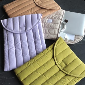 Op de afbeelding: Een verzameling gewatteerde laptoptassen in verschillende kleuren, waaronder lavendel, olijfgroen, bruin en beige. Elke tas heeft een klepsluiting en een gewatteerd ontwerp. Eén tas bevat een laptop en accessoires.