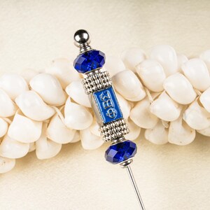 May include: A silver-toned decorative pin with a blue enamel center featuring Greek letters. The pin has a faceted blue crystal at the top and bottom, with a silver ball at the very top. The pin is set against a background of white seashells.
