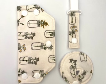 Könnte beinhalten: Ein Set Baby-Accessoires aus cremefarbenem Stoff mit einem botanischen Aufdruck von Gläsern mit Blumen. Das Set enthält eine Lätzchen, einen Schnullerclip und ein rundes Spucktuch, alle mit weißen Druckknöpfen.