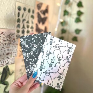 May include: Two small rectangular cards with dancing skeleton designs. One card is black with white skeletons, and the other is white with black skeletons. The cards are held in front of a wall with botanical prints and fairy lights.