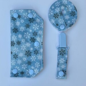 以下が含まれることがあります： 青と白の雪の結晶柄のベビー用アクセサリー3点セット。セットには、よだれかけ、歯固め、おしゃぶりクリップが含まれています。