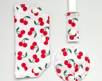 May include: A set of baby accessories featuring a white fabric with a red cherry pattern and green leaves. The set includes a pacifier clip, a round item, and a shaped item, all with white snap closures.