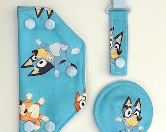 May include: A blue and brown fabric baby bib with a cartoon dog design, a matching blue fabric pacifier clip, and a blue fabric burp cloth with a cartoon dog design.