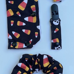 May include: A set of three Halloween-themed hair accessories featuring a black and orange candy corn pattern with a skull design. The set includes a hair scrunchie, a hair clip, and a pacifier clip.
