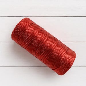 May include: A cylindrical spool of vibrant red cord featuring a tightly woven, braided texture with a subtle metallic sheen. The cord is wound in a continuous pattern, creating a uniform, dense surface that reflects light across its fibres.