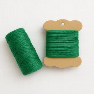 May include: A vibrant green yarn bundle positioned next to a light brown cardboard spool wrapped with the same green yarn. The spool features a scalloped edge design and a circular hole at the top for hanging or storage purposes.