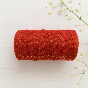 May include: A cylindrical spool of vibrant red cord featuring interwoven metallic gold threads. The cord is tightly wound into a compact, horizontal bundle, showcasing a consistent texture and a bright, reflective finish throughout the entire length.